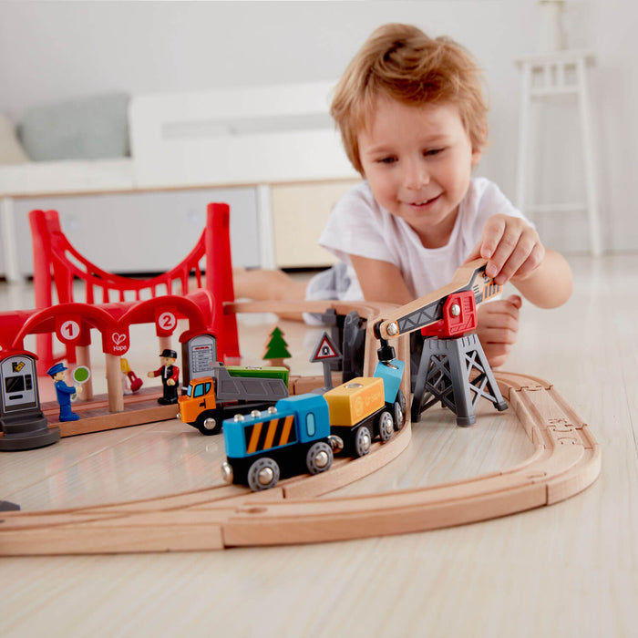 Child plays with a wooden train set featuring a red bridge, colorful train cars, and a crane on tracks.