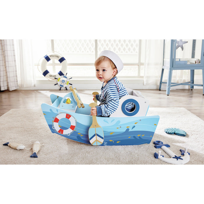 Toddler in a sailor hat sitting in a blue rocking boat toy with wave graphics, life ring detail, and a wooden oar.