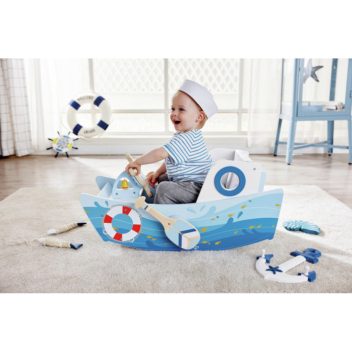 Toddler in a sailor hat sits in a blue wooden rocking boat with ocean wave graphics and a toy oar on a soft rug.