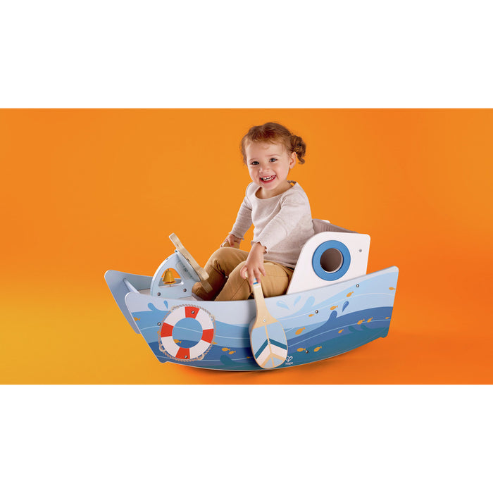Child sitting in a blue wooden rocking boat with a steering wheel and oar against a solid orange background.