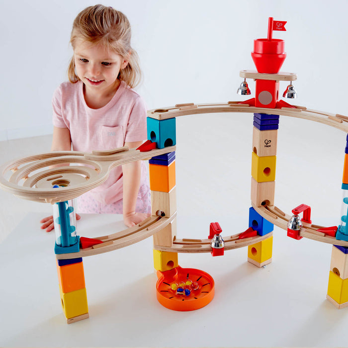A girl plays with a wooden marble run featuring colorful blocks, curved tracks, silver bells, and a red funnel flag.
