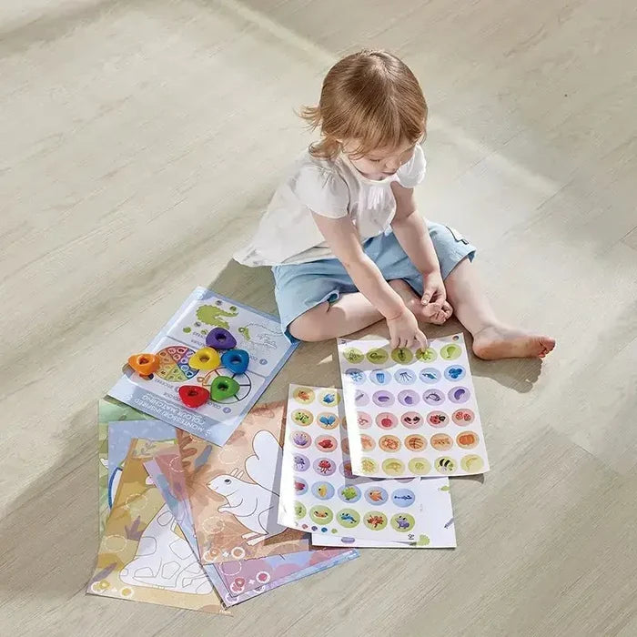 A toddler uses colorful triangular crayons with illustrated activity sheets on a light wood floor.