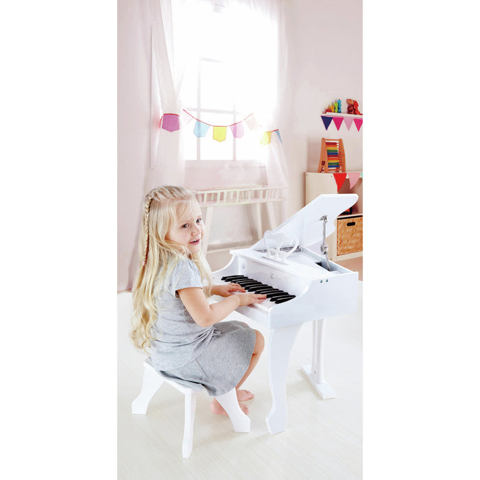 Young girl playing a white toy grand piano with an open lid and a matching stool in a bright room.
