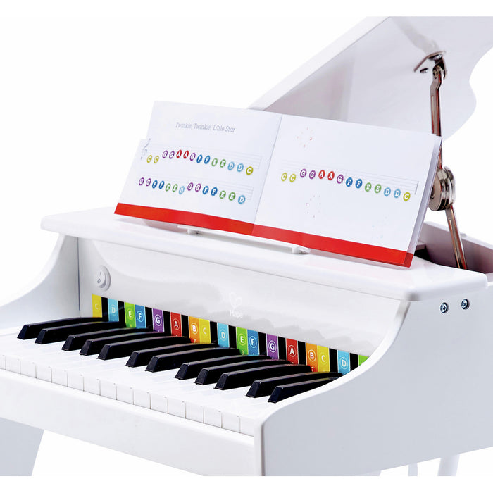 White toy grand piano with color-coded key labels and an open music book on the stand.