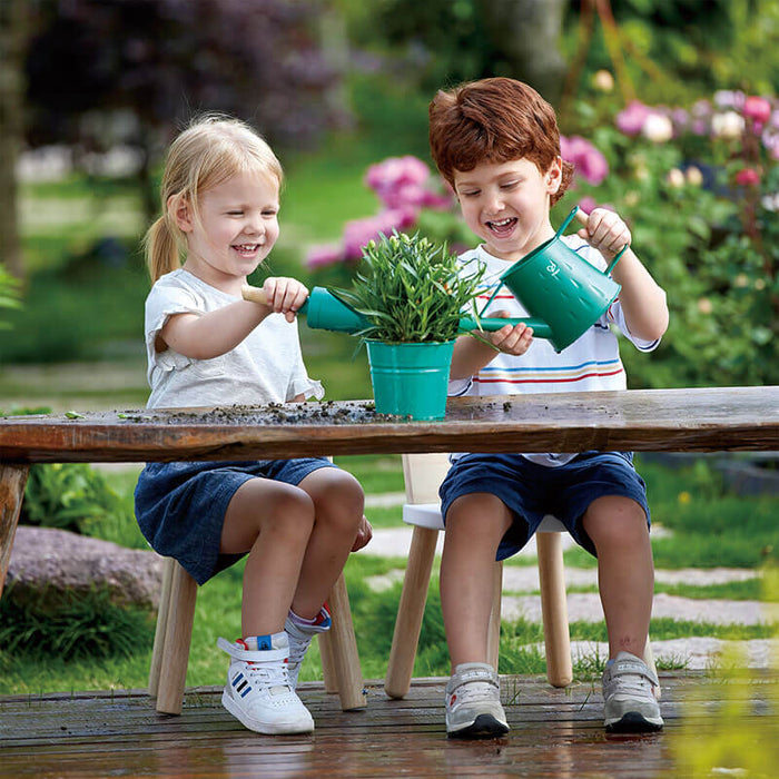 Two children playing with a teal toy watering can and matching pot at a wooden garden table.