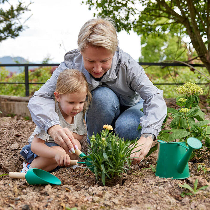 Woman and child use green garden tools with wooden handles and a watering can to plant a small flower in soil.