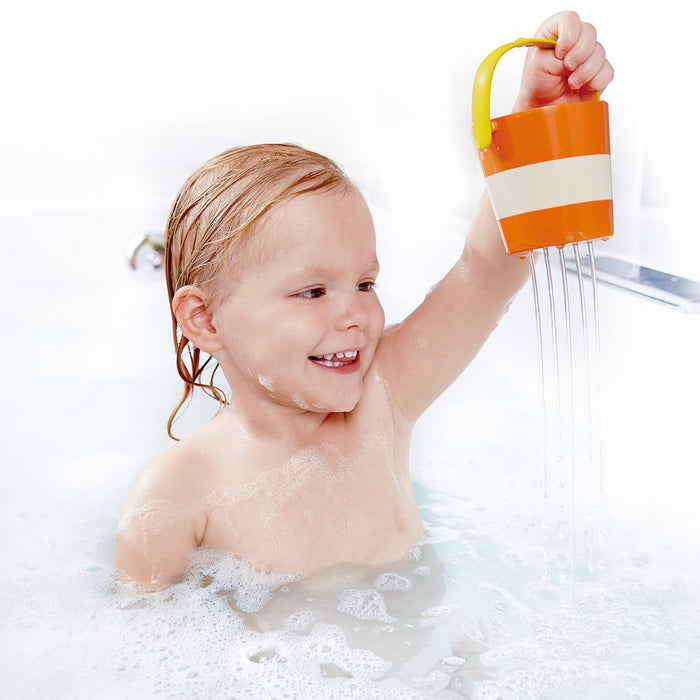 Smiling toddler in a bubble bath holding an orange striped bucket with water raining from holes in the bottom.