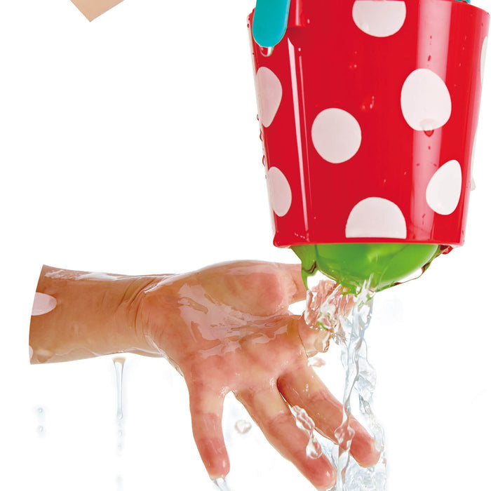 Red bucket with white polka dots pouring water from its green base onto a child's hand.