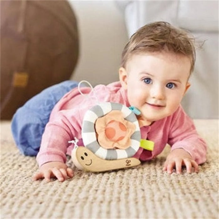 Baby lying on a rug with a wooden snail toy featuring a striped grey and white plush shell and a smiling face.
