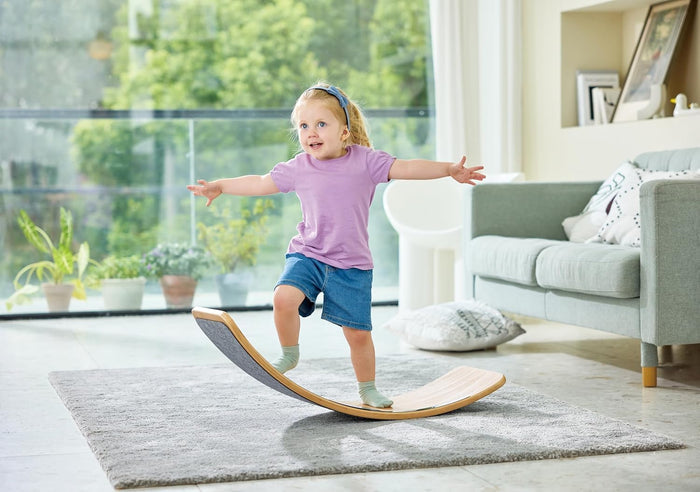 A young girl balancing on a curved wooden rocker board with gray felt grip in a bright living room.