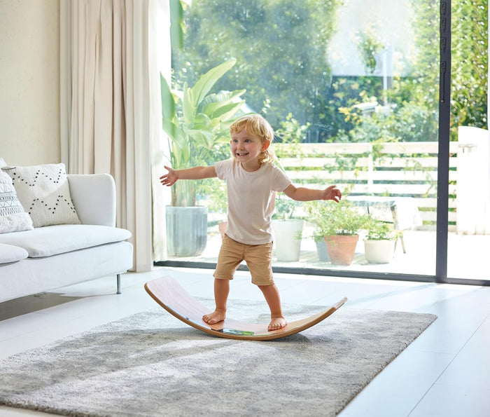Child balancing on a curved wooden board with a glowing green sensor on a grey rug in a bright living room.