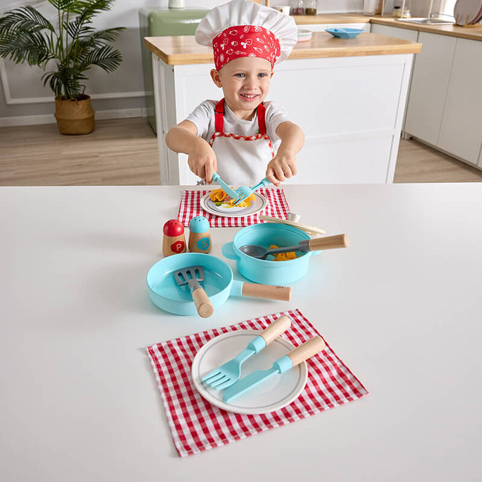 Child in a chef hat playing with a blue toy cookware set, wooden utensils, and red checkered placemats.