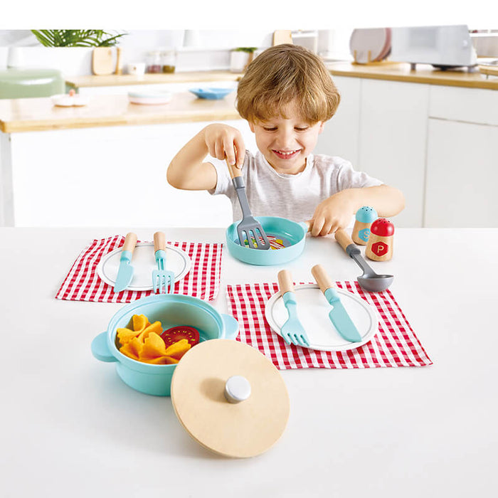 Child playing with light blue and wood toy cooking set including a pot, pan, and cutlery on red checkered napkins.