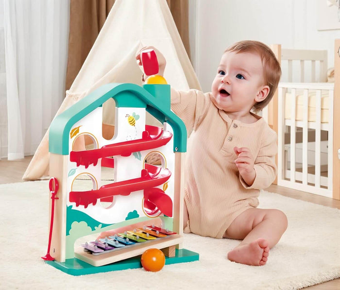 Teal wooden house ball run with red ramps, rainbow xylophone, and orange balls, shown with a baby playing.