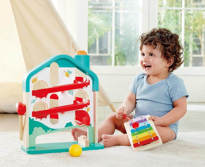 Toddler playing with a teal house-shaped wooden ball run with red tracks and a small rainbow xylophone.