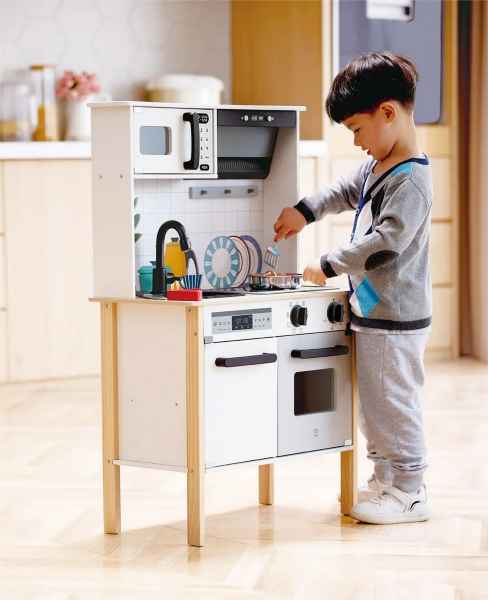 Child playing with a white and wood toy kitchen featuring a microwave, sink, stove, and oven.