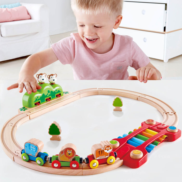 Toddler playing with a wooden railway set featuring animal cars, a monkey tunnel, and a colorful xylophone track.