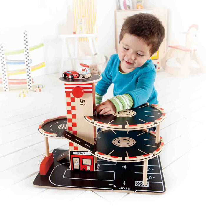 Toddler plays with a multi-level wooden toy garage featuring black decks, red ramps, and a rooftop helipad with toy cars.