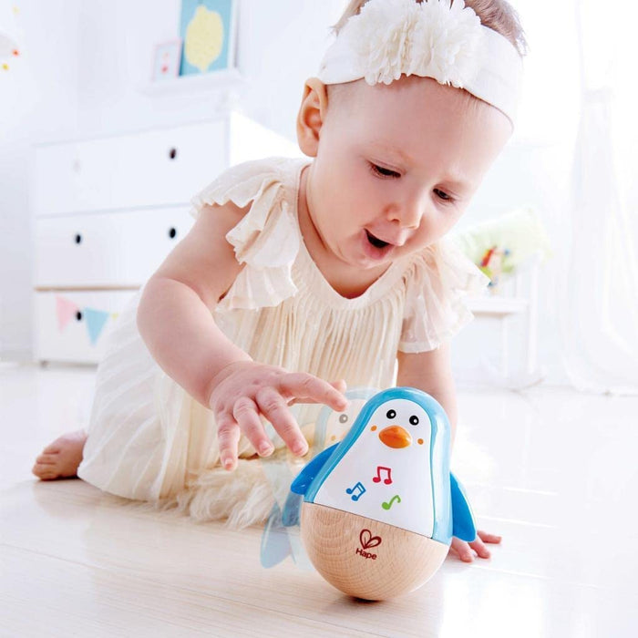 Baby girl reaches for a blue and white penguin wobbler toy with a rounded wooden base and musical note icons.