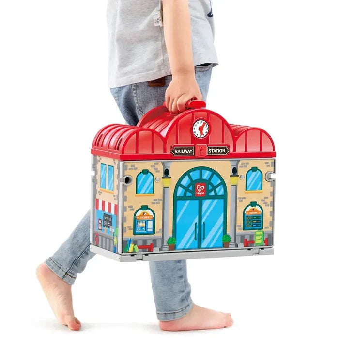 Child carrying a red and tan toy railway station with a handle, arched blue doors, and a clock on the roof.