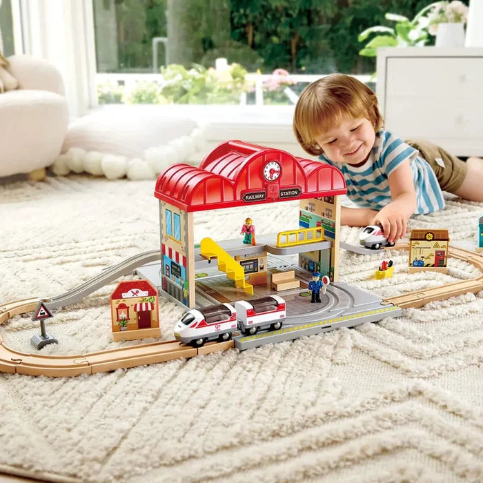 Child playing with a red two-story toy station, wooden tracks, and white bullet trains on a plush white rug.