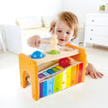 Toddler hits colorful balls through a yellow wooden bench onto a rainbow slide-out xylophone with a wooden mallet.