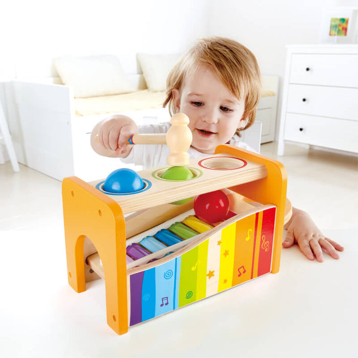 Toddler hits colorful balls through a yellow wooden bench onto a rainbow slide-out xylophone with a wooden mallet.