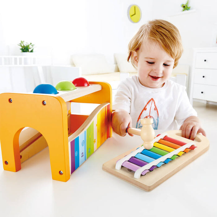 Child using a wooden mallet to tap a colorful xylophone next to a yellow pound bench with red, blue, and green balls.