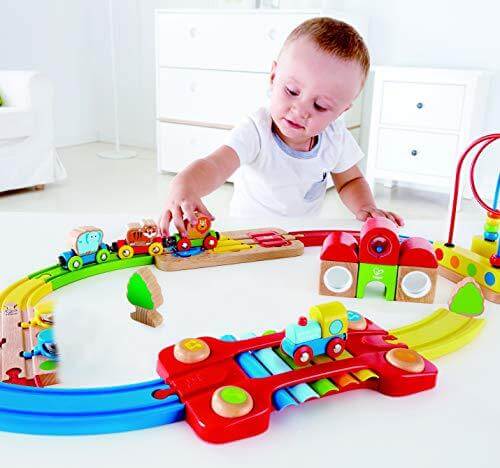 Toddler playing with a vibrant wooden train set with animal-themed cars and a rainbow xylophone bridge track.
