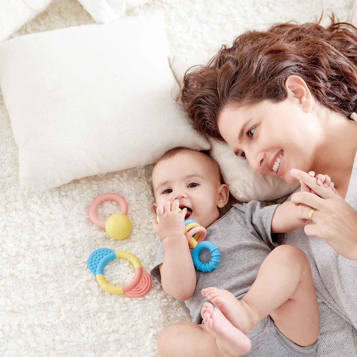 Baby and mother lying on a white rug with three colorful teething rattles in pastel blue, yellow, and pink.