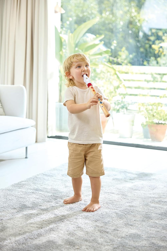 A young child plays a light wood recorder with colorful dots and a white mouthpiece, standing on a grey rug.