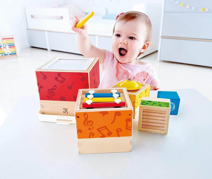Excited baby playing with colorful wooden stacking boxes featuring a xylophone, drum, and bell.