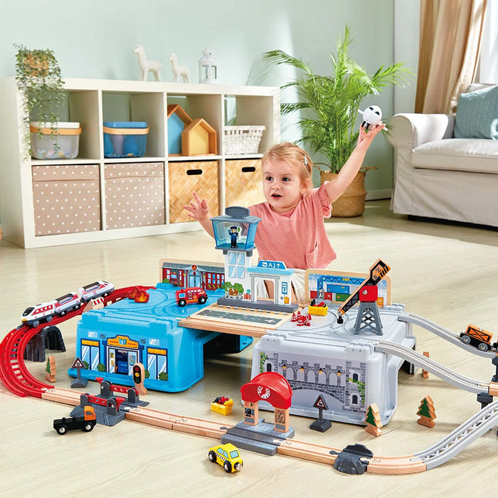 Toddler playing with a toy city set featuring wooden tracks, blue and gray buckets, a red crane, and colorful vehicles.