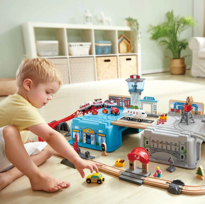 Child plays with a multi-level toy cityscape with wooden tracks, a blue bucket base, a red train, and an airplane.
