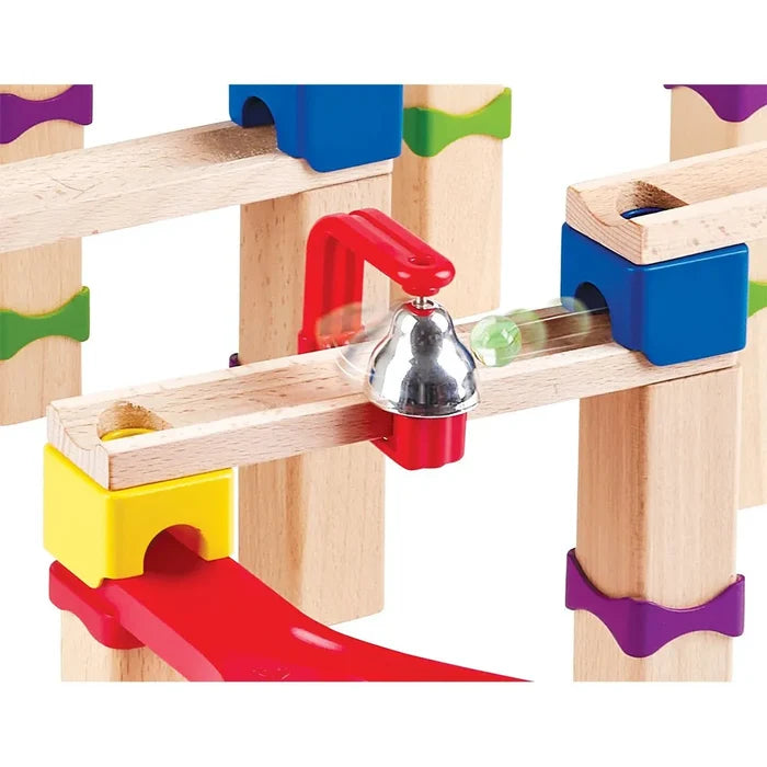 Close-up of a wooden marble run with colorful supports, a silver bell, and a green marble rolling down a track.