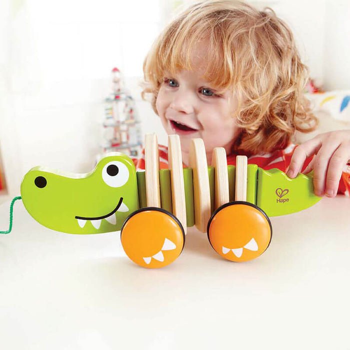 Green wooden crocodile pull toy with orange wheels and a slatted body, shown with a smiling toddler.