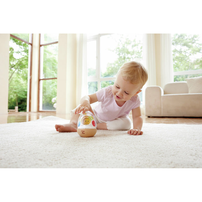 Toddler playing with a wooden-based wobble toy with a clear dome and colorful interior on a white rug.