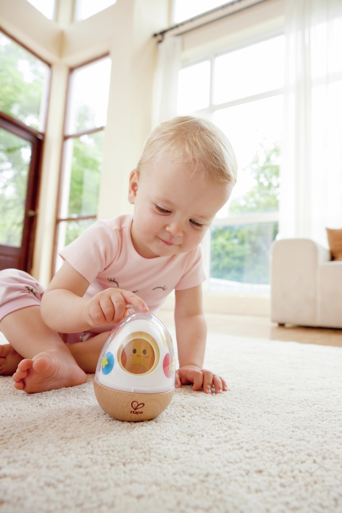 Toddler playing with an egg-shaped wooden wobble toy with a yellow chick inside on a plush rug.