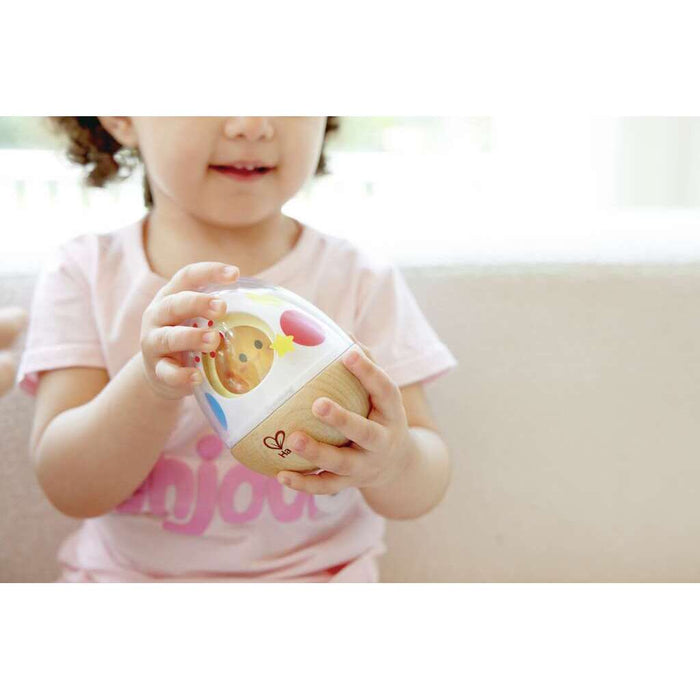 Toddler holding a wooden egg-shaped wobble toy with a clear plastic top, colorful patterns, and a red heart logo.