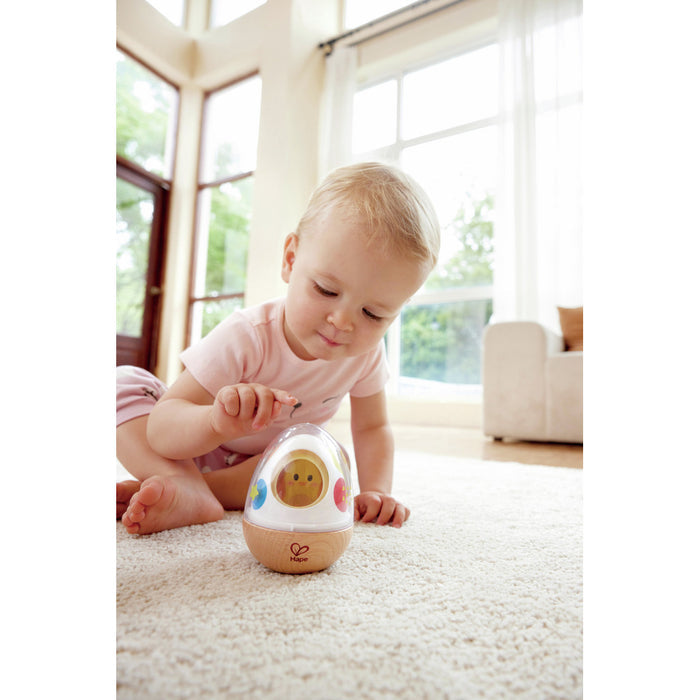 Toddler playing with an egg-shaped wooden wobble toy with a clear top and yellow chick inside on a white rug.