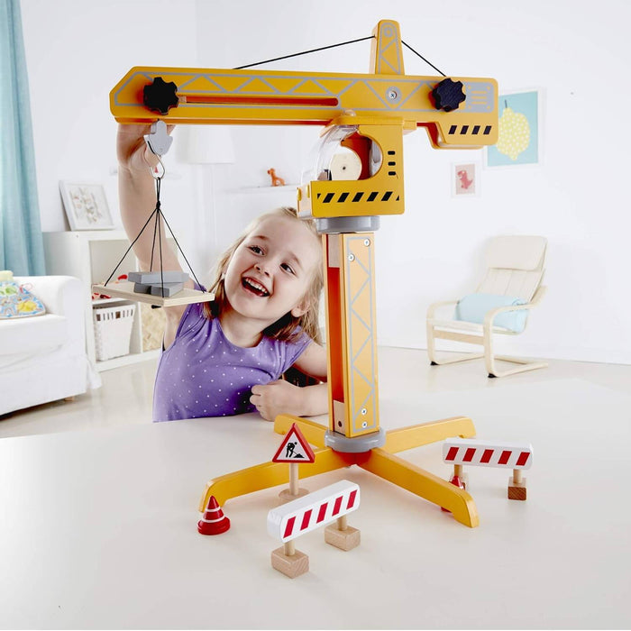 Smiling girl playing with a yellow wooden crane toy featuring a pulley, lifting platform, and construction signs.
