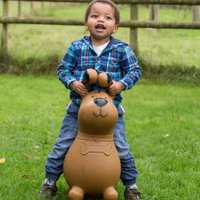 Young child riding a brown inflatable dog-shaped hopper toy with black ears and a bone collar on a grassy lawn.