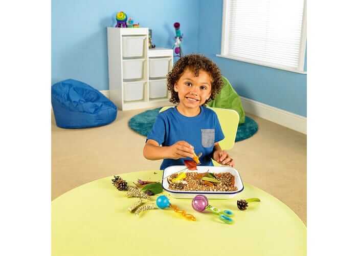 A boy sits at a yellow table using colorful scoops and tweezers to sort natural materials from a tray.