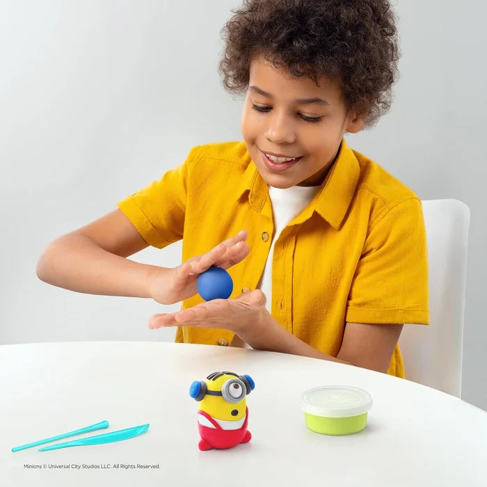 A child rolls blue clay next to a yellow Minion figurine, blue sculpting tools, and a green tub on a white table.