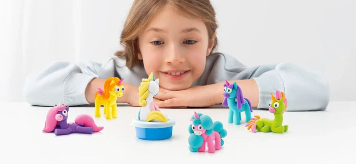 A smiling girl looks at six colorful, stylized clay unicorns in bright shades of yellow, purple, blue, and green.