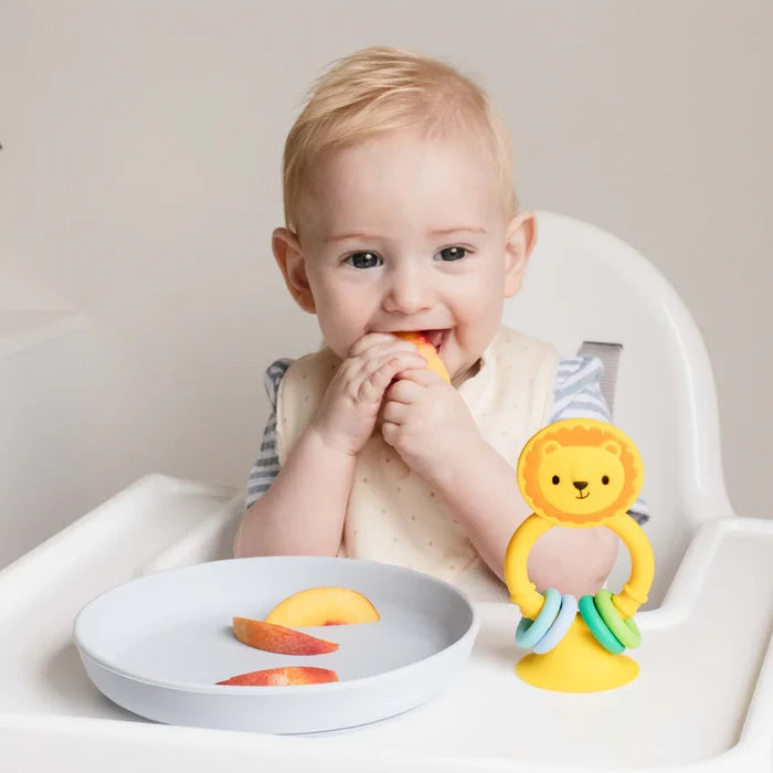 Yellow silicone lion toy with a suction base and colorful rings on a high chair tray next to a baby eating fruit.