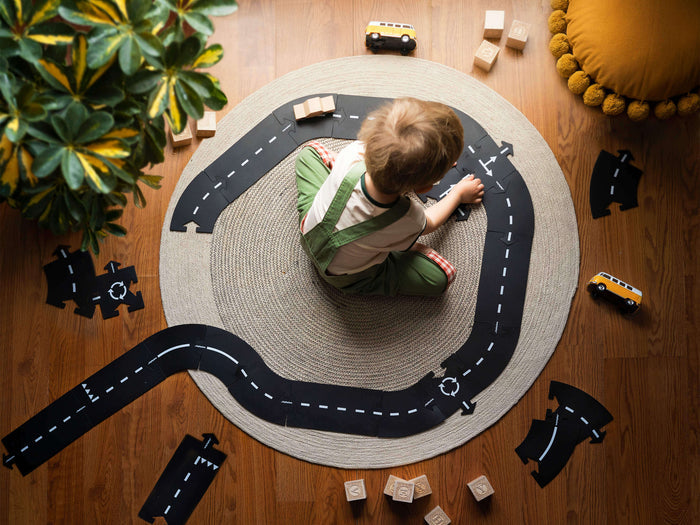 A child assembles flexible black toy road pieces with white markings into a track on a round rug.
