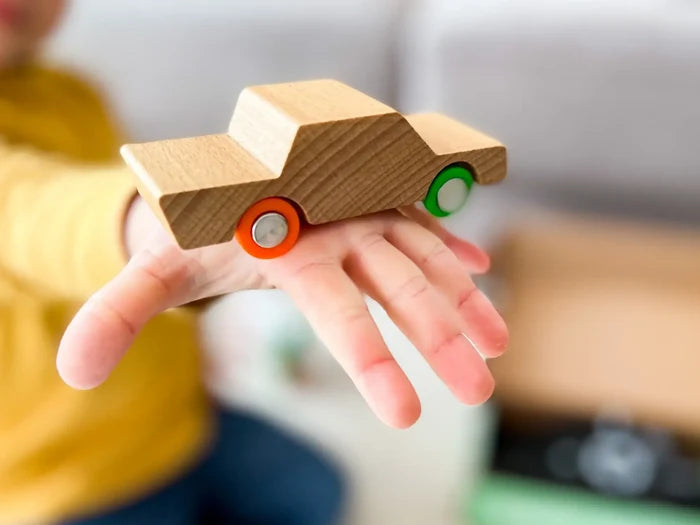 Minimalist wooden toy car with one orange wheel and one green wheel, balanced on a child's hand.