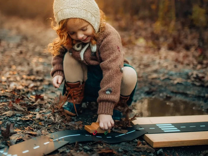 A young girl playing with flexible black toy road tracks and a small wooden car on a leafy outdoor path.