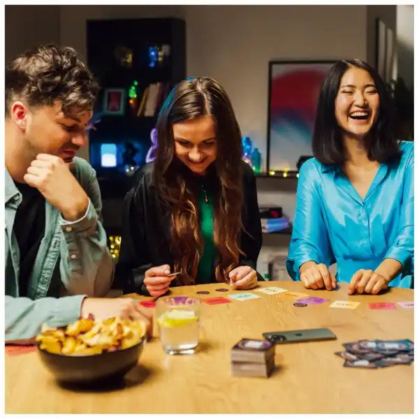 Three friends laughing while playing a music game with colorful square cards, round tokens, and a smartphone.